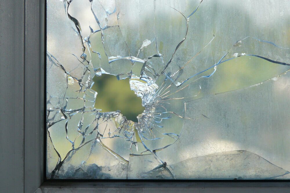 Hole in the window glass by a bullet shot during war shooting. Cracks spreading around the hole. Green leaves of trees blurred by the glass. Dirty window frame. View of a street from the inside.