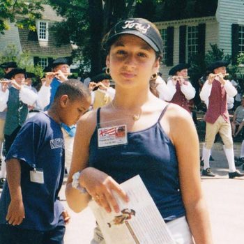 Girl holding a paper, standing in front of a marching band in colonial attire, outdoors with trees and historic buildings in the background.