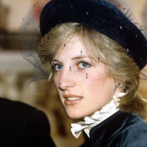 A person, wearing a dark blue veiled hat, gazes sideways in a formal indoor setting.
