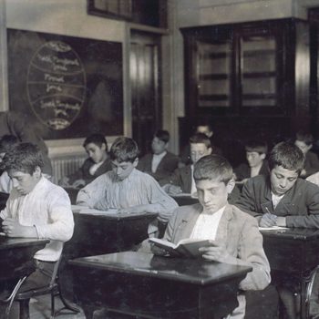 Students attentively read and write at desks in a classroom. A teacher assists a student; chalkboard diagrams are visible in the background.