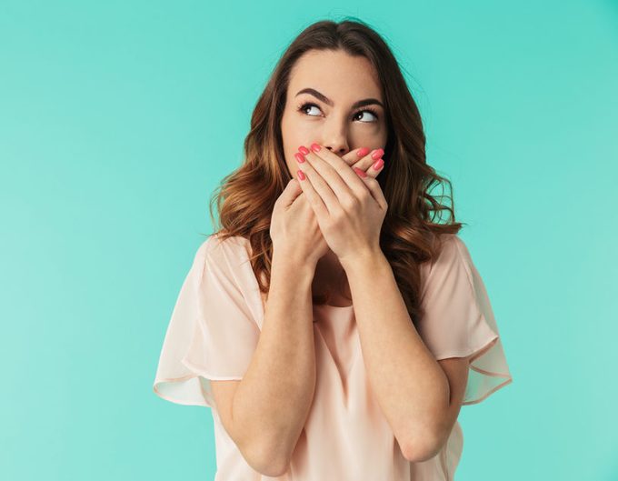 Woman covering mouth with hands, looking to the side, against a solid teal background.