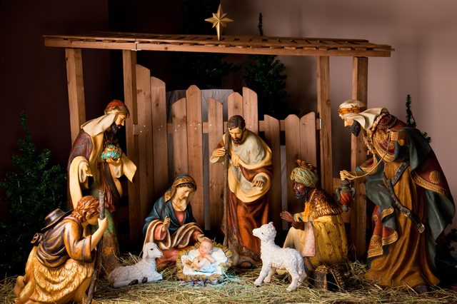 Figurines surround a baby in a manger, depicting a nativity scene, under a wooden shelter with straw and lambs, surrounded by trees.
