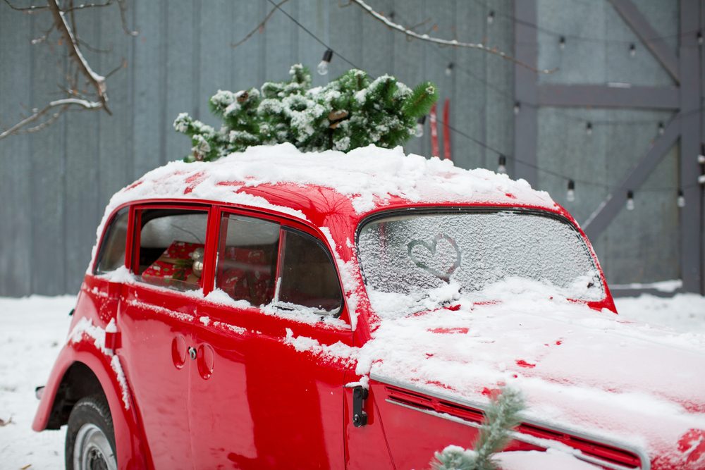 A red car; covered in snow; parked outside with a tree on the roof and heart shape on windshield.