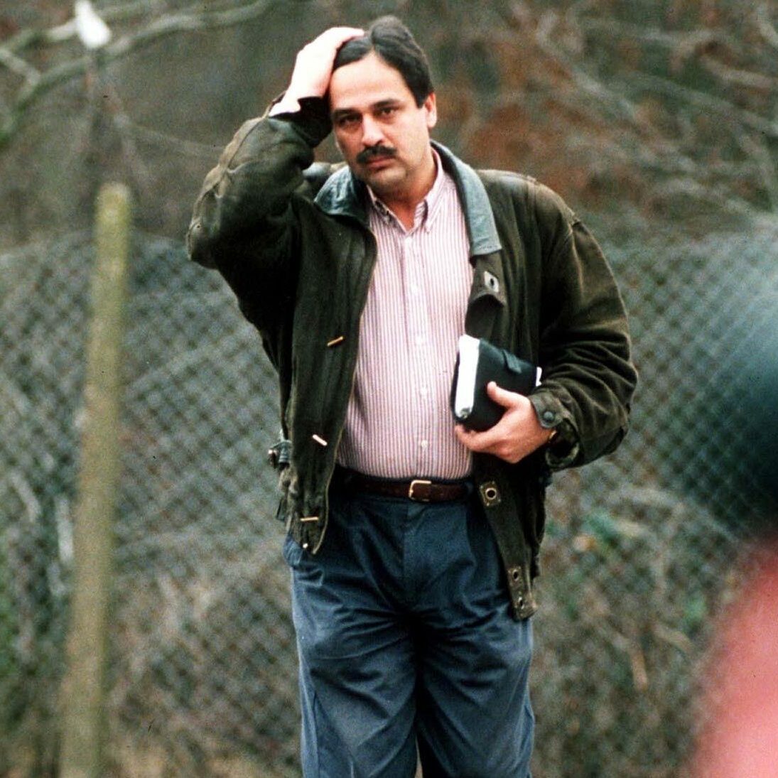 Man walking outdoors, holding a book and adjusting his hair, with a chain-link fence and leaf-covered ground in the background.