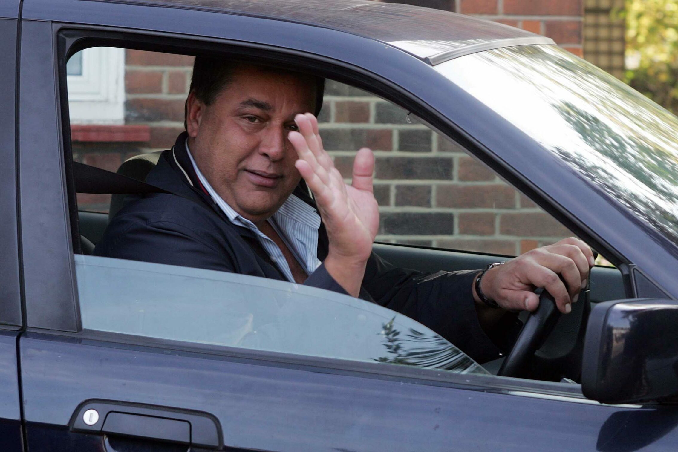 Person driving a dark car, waving through an open window, parked outside a brick building.