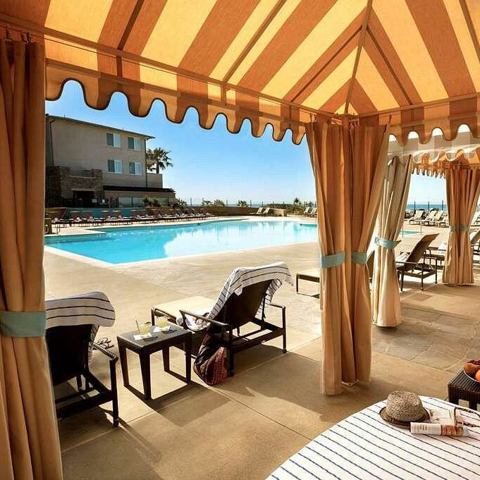 Lounge chairs rest under cabanas beside a pool, amid a resort setting with palm trees and buildings in the background.