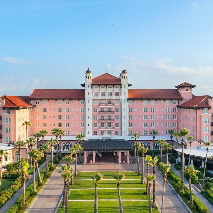 Pink hotel stands surrounded by palm trees, featuring a red roof and lush gardens, under a clear blue sky.