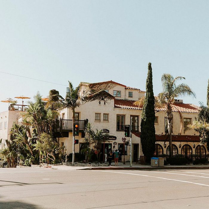 A Spanish-style hotel sits on a sunny street corner, surrounded by palm trees. People walk near the entrance, and traffic lights are visible.