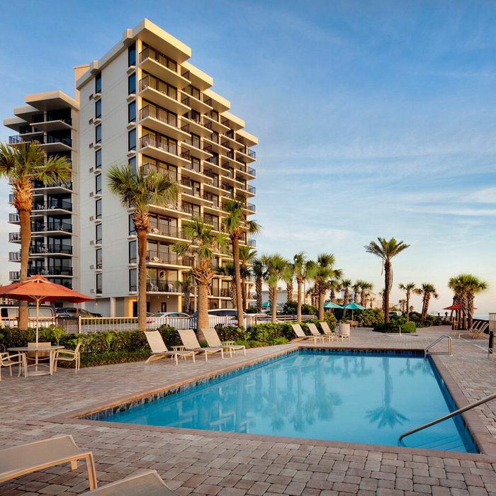 A swimming pool sits surrounded by lounge chairs, near a tall building, with palm trees and an ocean view under a clear sky.
