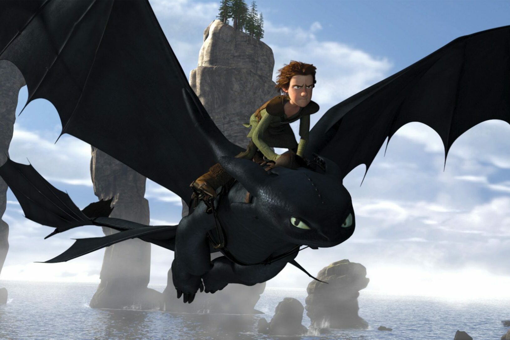 A dragon with a rider flies between towering rock formations over a misty sea, under a clear blue sky.