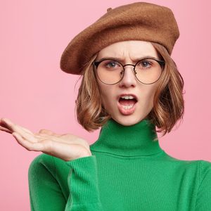 Person wearing glasses and a beret gestures with hand, expressing surprise, set against a plain pink background; wearing a green turtleneck sweater.
