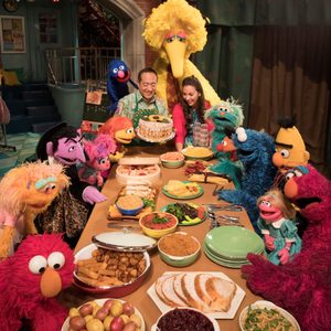 Puppets and two people gather around a table filled with diverse foods, celebrating in a warmly lit, inviting room.