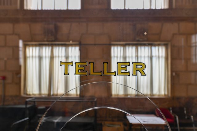 "TELLER" text appears on glass, revealing a bank interior with high windows and curtains.