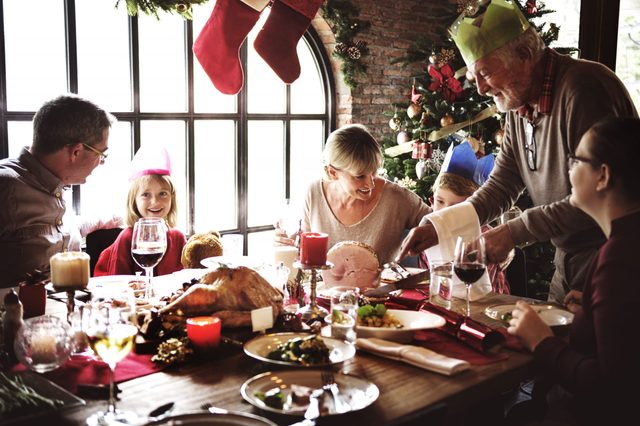 Family sharing a festive meal, wearing paper hats, around a decorated Christmas tree and dining table with candles, food, and wine.
