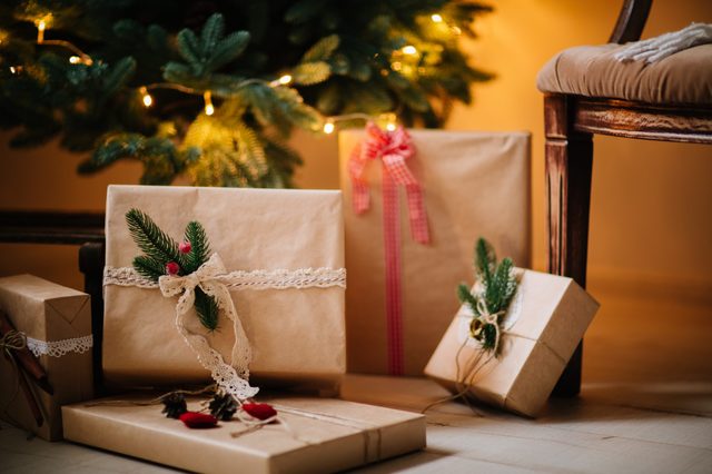 Presents wrapped in brown paper with lace and pine accents rest under a Christmas tree adorned with warm lights, next to a wooden chair.