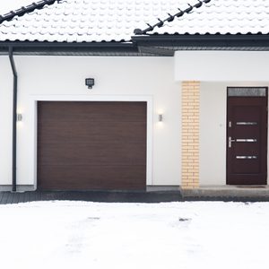 A snow-covered house with a dark garage door and front door, set in a wintry landscape with bare trees and a clear path.
