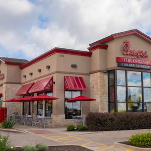 Restaurant building with red awnings and umbrellas, surrounded by greenery, in a bright outdoor setting. Sign reads 