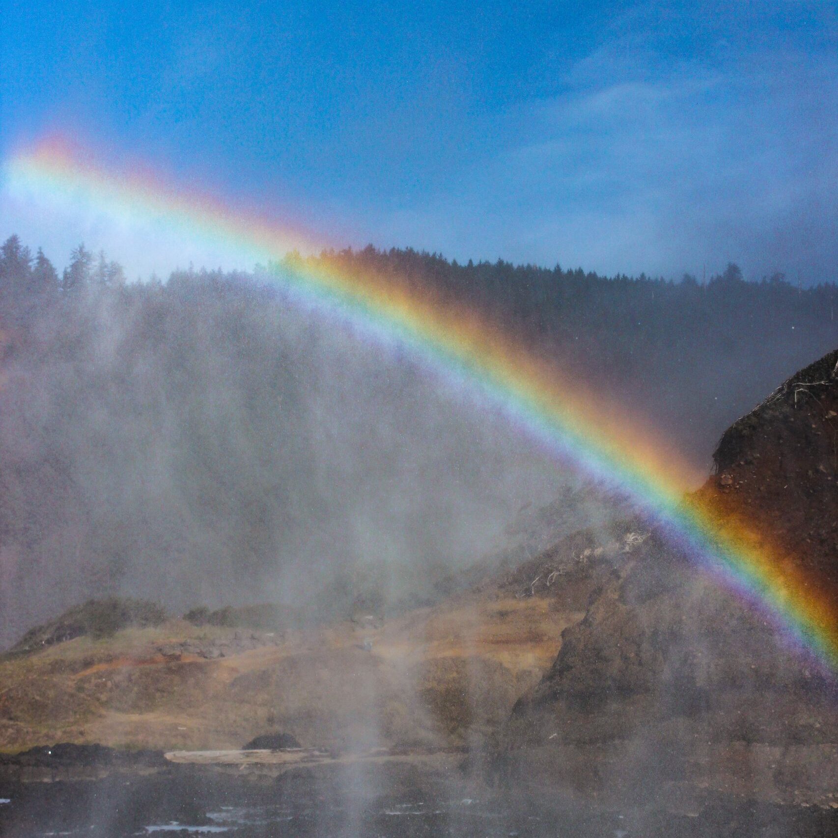 Rainbow arches brightly over misty rugged coastline, with dark rocks and foamy waves beneath a clear blue sky.