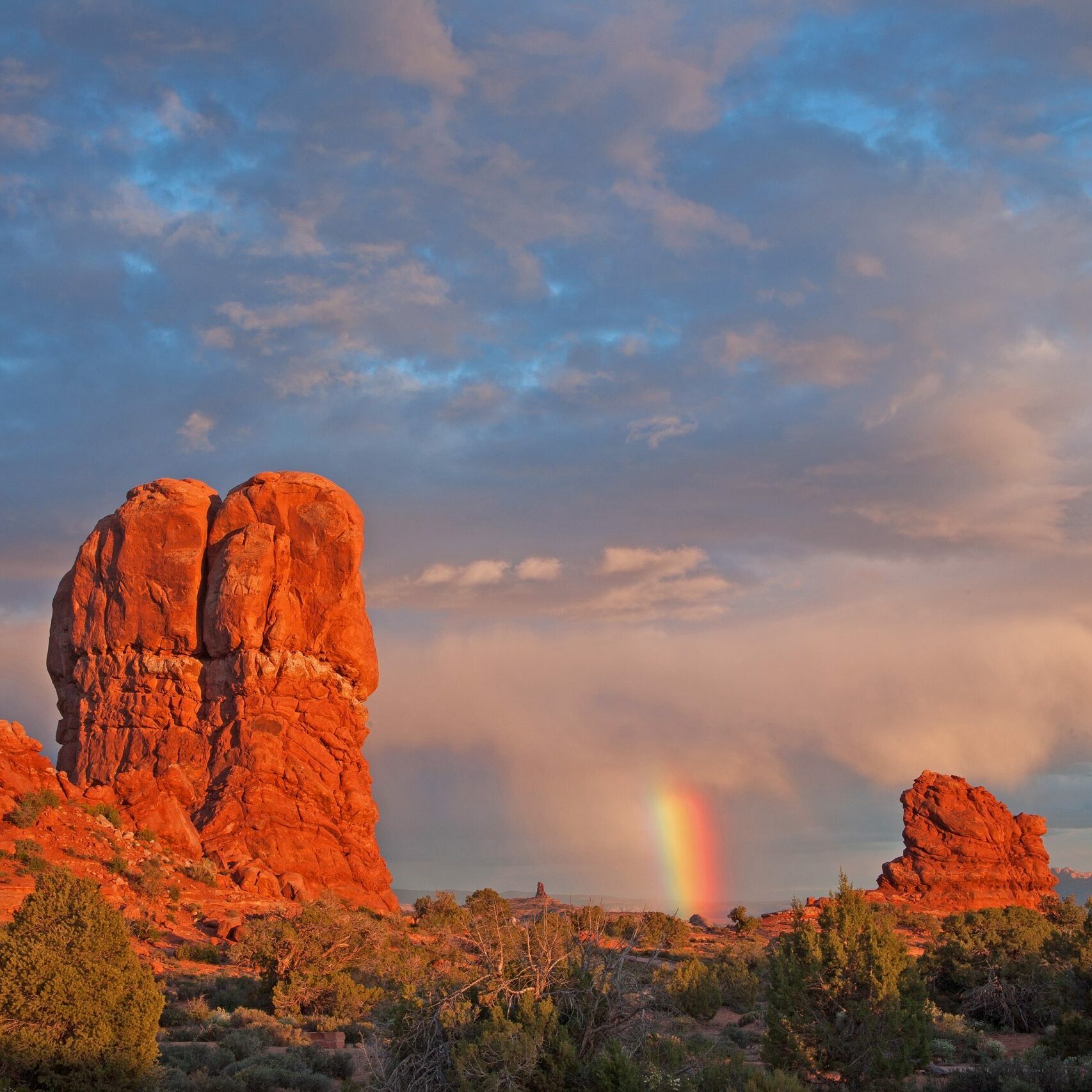 Red rock formations tower under vivid clouds; a rainbow arcs in the distance, enhancing the dramatic desert landscape.