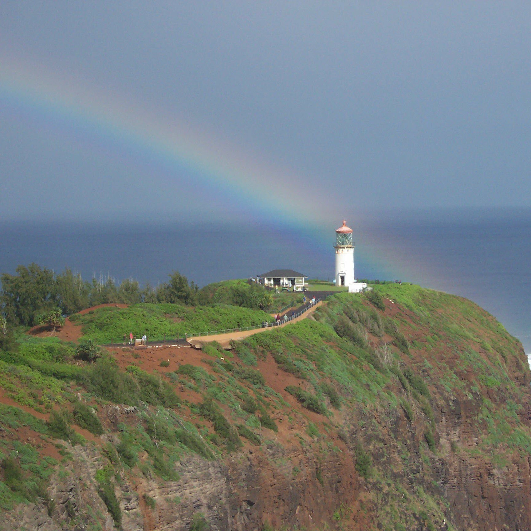 Lighthouse stands on lush cliff, overlooking the sea; a vibrant rainbow arches across the sky. Waves crash against the rocky shoreline below.