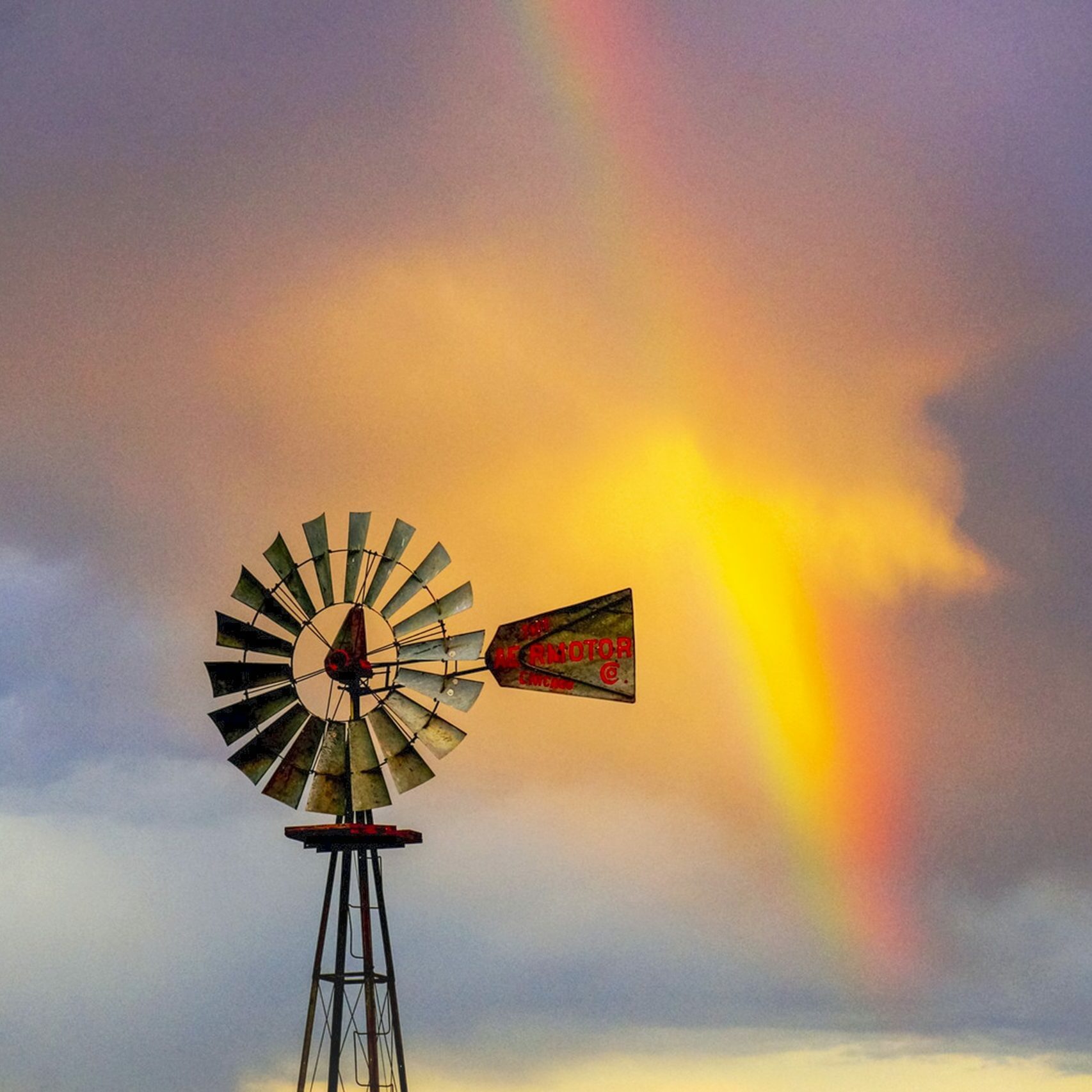 A windmill stands against a dramatic sky with vibrant rainbow colors illuminating the clouds.