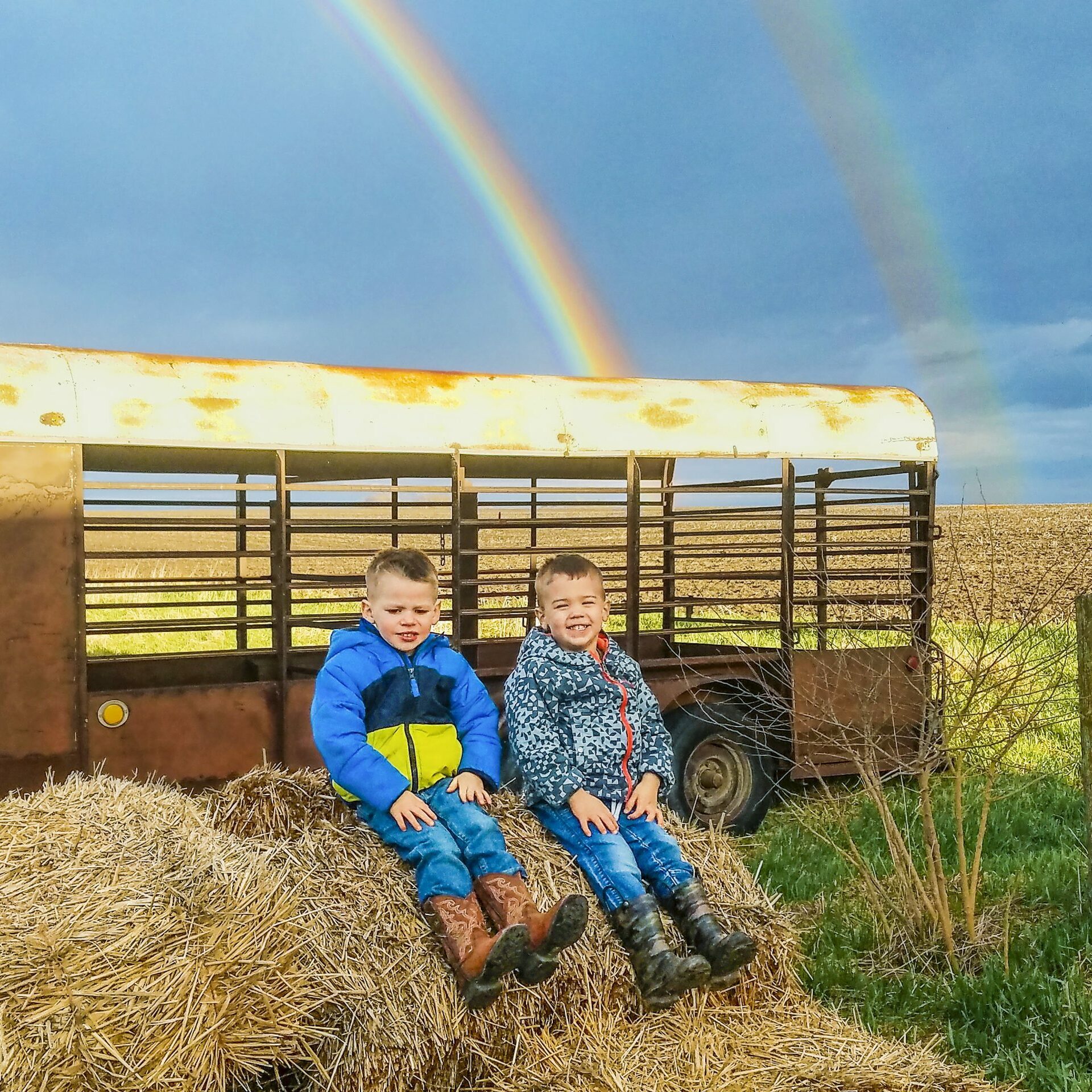 Two children sit smiling on hay bales, with a rusty trailer and a vibrant double rainbow in a rural landscape.