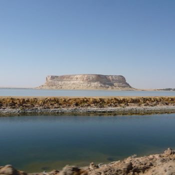 A mesa stands motionless surrounded by desert and a calm body of water under a clear blue sky.