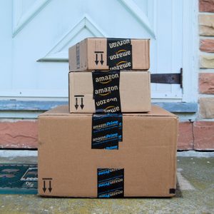 Three stacked Amazon packages sit on a doorstep in front of a white door, surrounded by a brick wall.