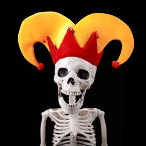 A smiling skeleton wears a colorful jester hat against a plain black background.