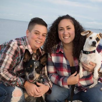 Two people smile, holding two dogs, seated by a calm waterfront. They wear plaid shirts.