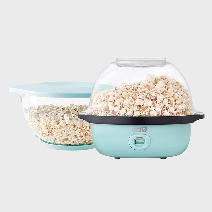 Popcorn maker popping popcorn, sitting on a plain surface next to a transparent bowl filled with popcorn.