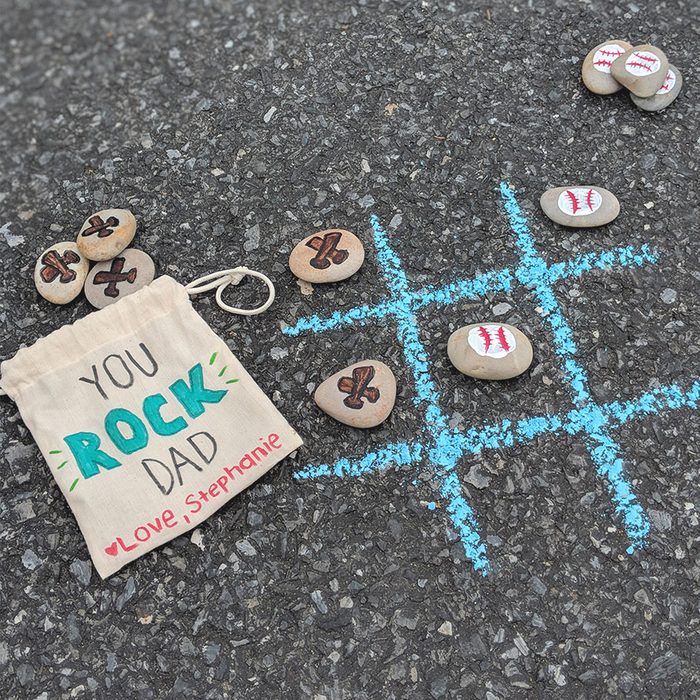 Stones painted with Xs and baseballs placed on a chalk tic-tac-toe grid on asphalt. A pouch reads 