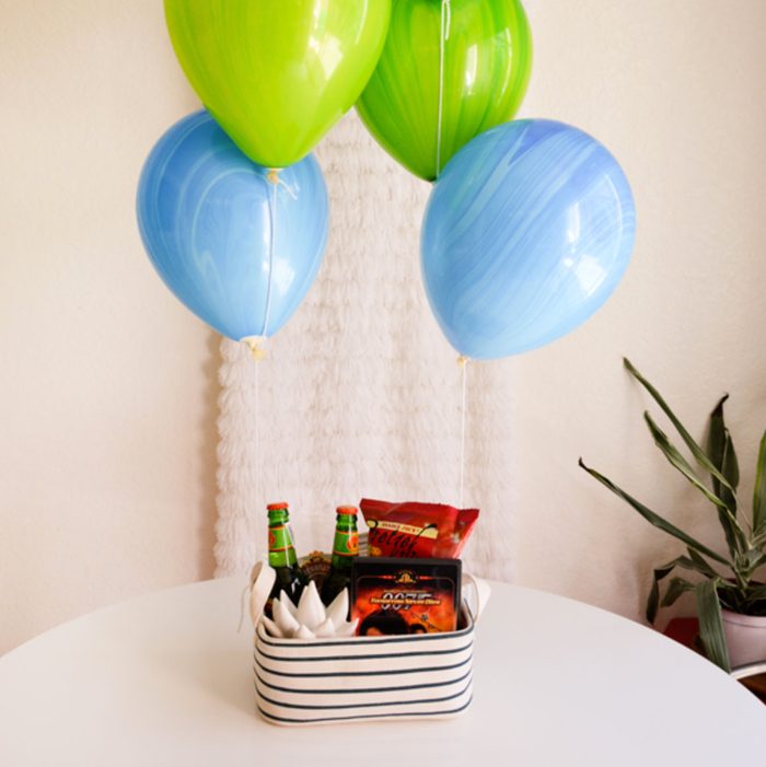 Gift basket with snacks and drinks sits on a table; balloons float above it in a room with plants.