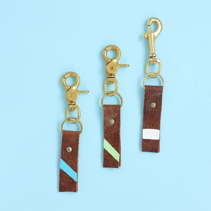 Three brown leather keychains with brass clasps lie on a light blue surface, each featuring different colored diagonal stripes.