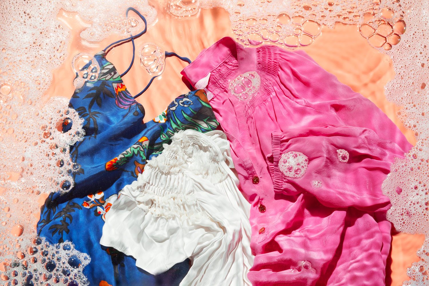 Clothes soak in soapy water, featuring a blue floral dress, white blouse, and pink shirt. The scene is filled with foamy bubbles.