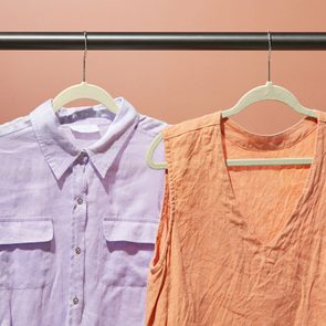 Shirts hang on a rod; a purple button-down with pockets and an orange sleeveless top, set against a warm, peach-colored background.