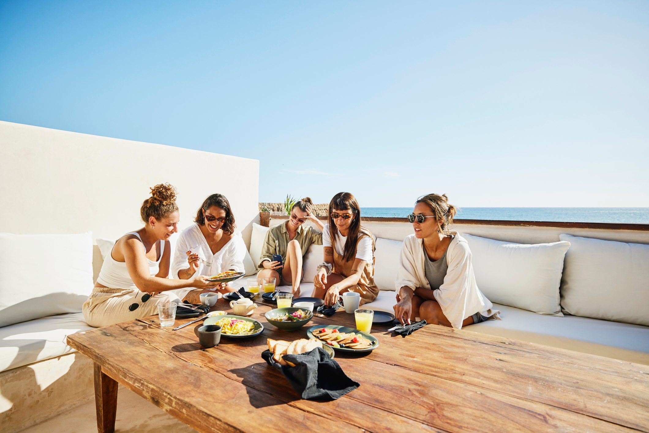 Wide shot of female friends sharing breakfast on deck of luxury suite at tropical resort