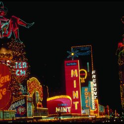Neon signs illuminate, advertising casinos and hotels at night, with colorful lights and a large cowgirl figure atop 