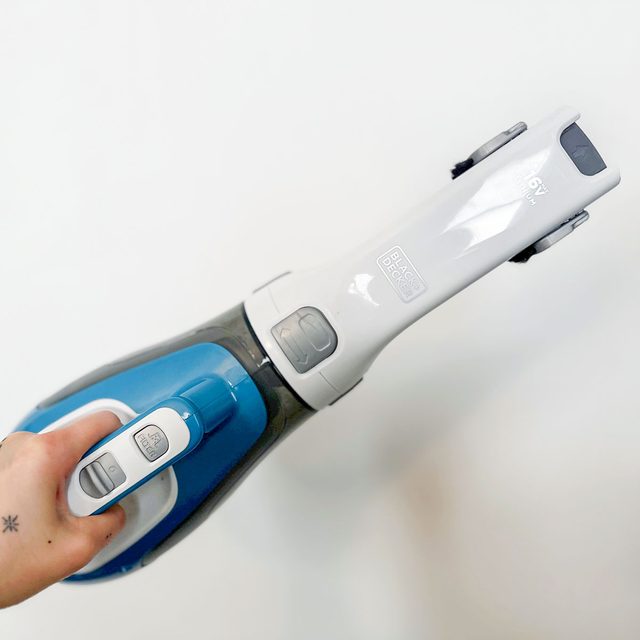 Hand holds blue and white handheld vacuum, operating in a plain, white background. Text on vacuum: 