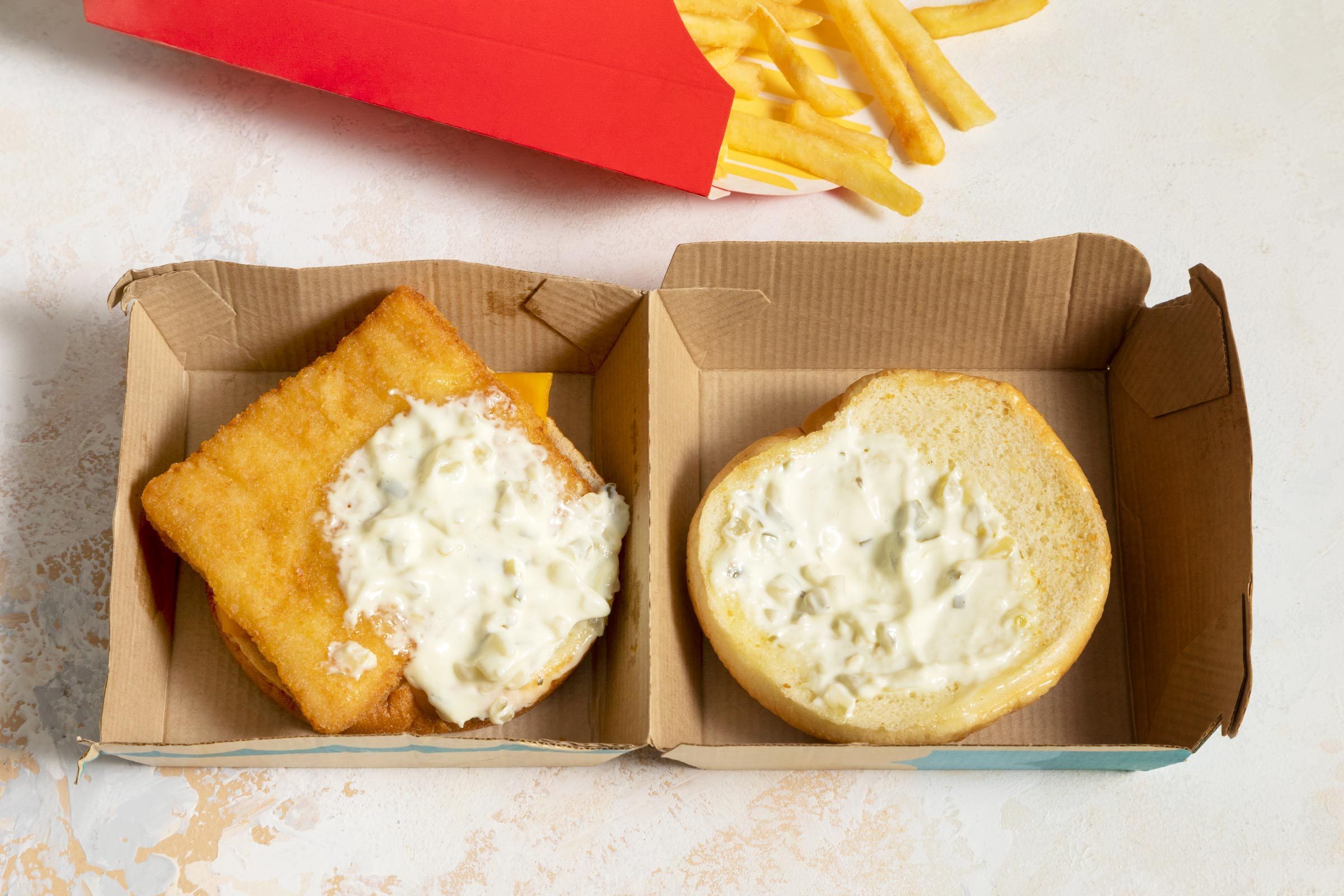 Fish sandwich with tartar sauce opened in a cardboard box; fries spill from a red container nearby on a light-colored surface.