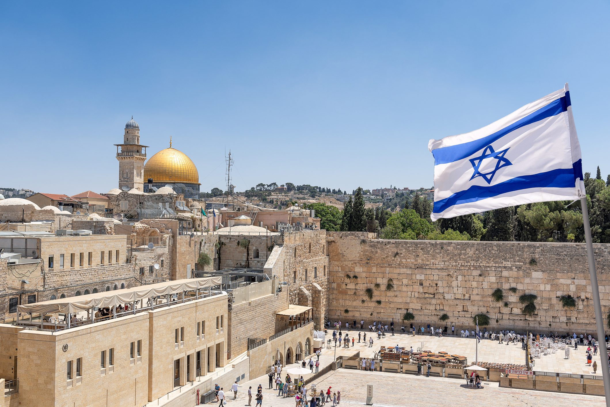 Israeli flag flutters above the Western Wall, with the golden Dome of the Rock and Jerusalem's historic architecture in the background under a clear sky.