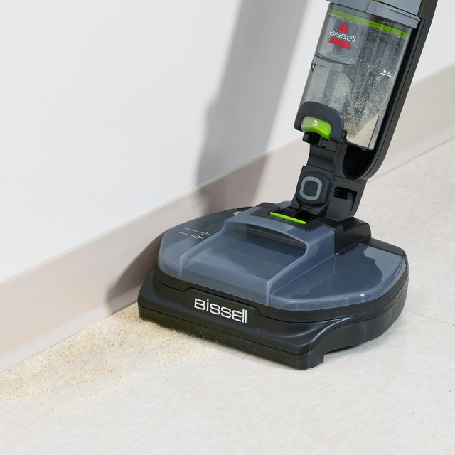 Vacuum cleaner suctioning dust along baseboard, on a tiled floor. The brand 