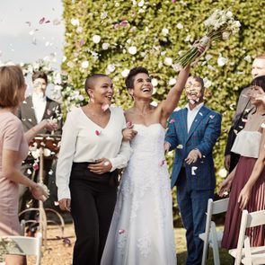 A bride raises a bouquet joyfully, surrounded by smiling guests tossing flower petals, in an outdoor wedding setting with abundant greenery.