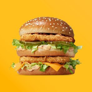A double chicken sandwich with lettuce, cheese, and sauce rests on a sesame seed bun against a bright yellow background.