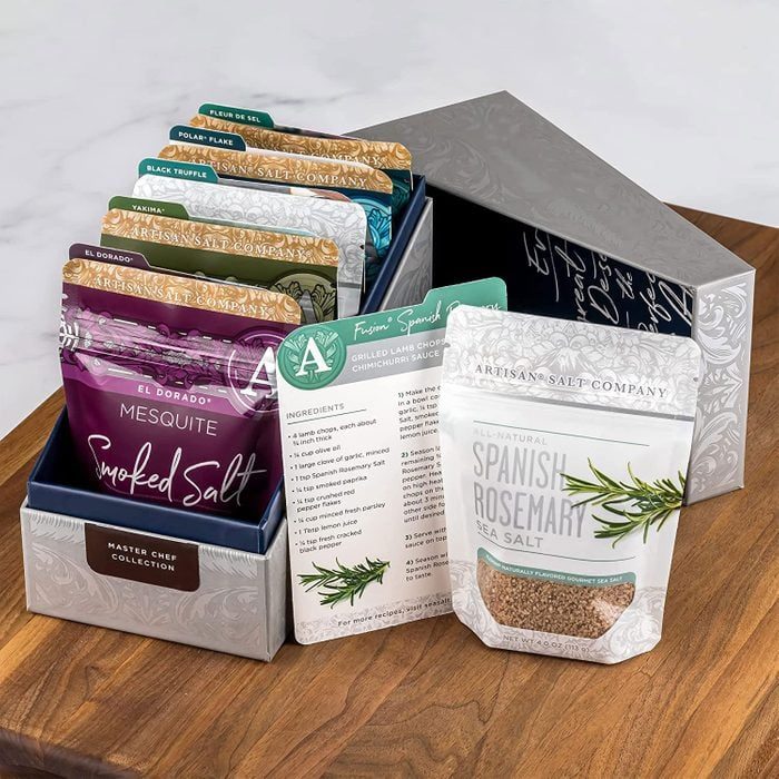 Packets of flavored salts displayed upright in an open decorative box on a wooden surface, accompanied by an informational card detailing ingredients and uses.
