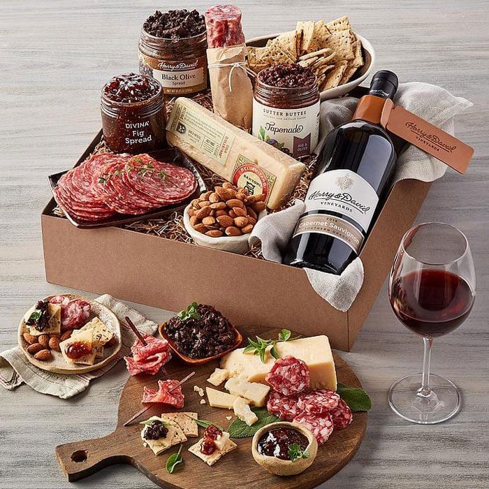 Gourmet gift box contains sliced meats, cheeses, nuts, and spreads, paired with a wine bottle, on a table next to a glass of wine.