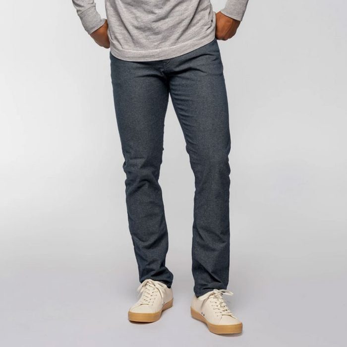 Person wears grey sweater and blue jeans, standing with hands in pockets, in a neutral indoor setting. Cream sneakers complete the casual attire.
