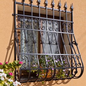 Iron window grate holds flowering plants outside closed window on a sunny orange wall beside a wooden door.