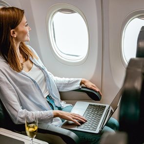 A woman sits in an airplane seat by the window, looking outside. She has a laptop on her lap and a glass of wine on the tray next to her. The cabin appears calm and well-lit.