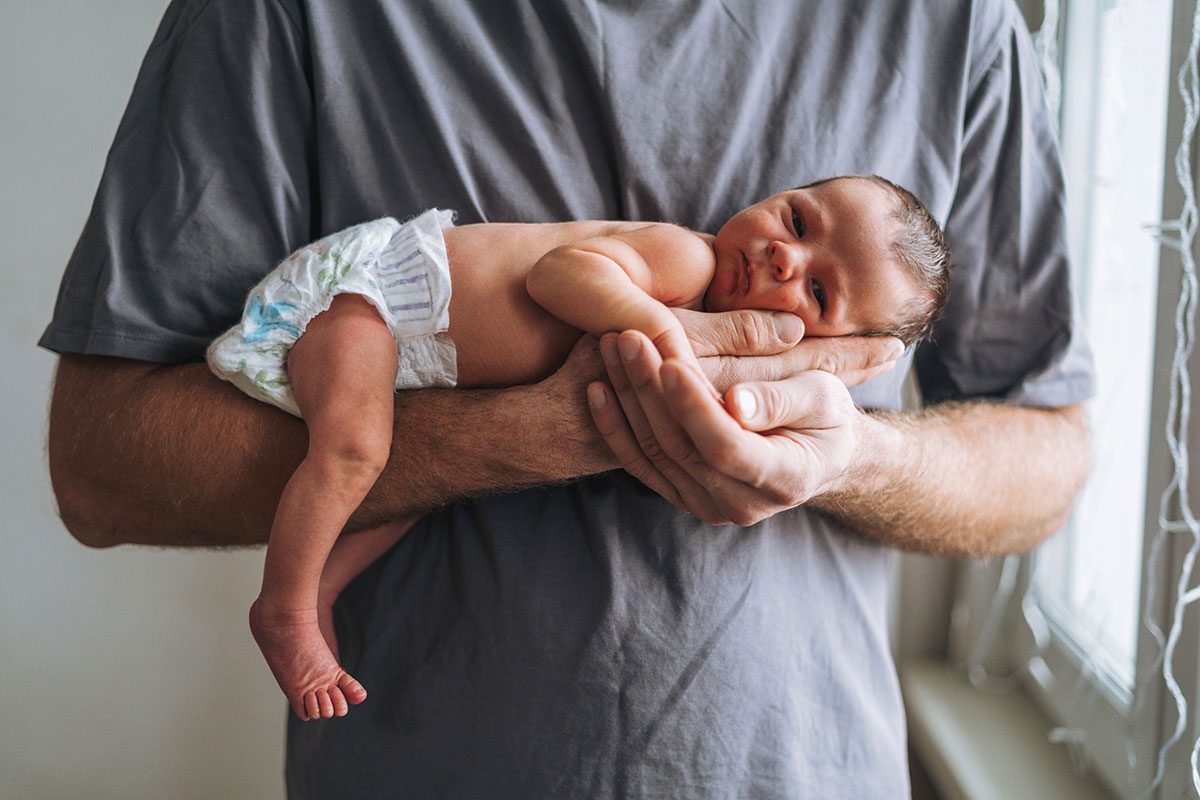 Young father with 2 weeks newborn baby boy in his hands near window at home